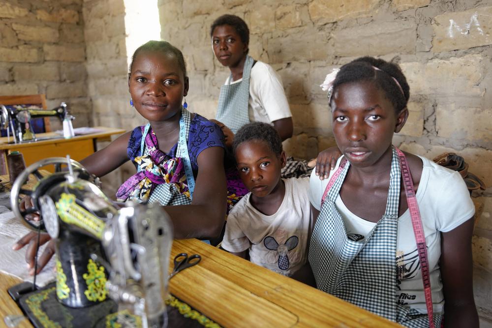 Quatre jeunes filles africaines, dont une est assise à une machine à coudre, les trois autres debout à côté delle, toutes souriantes.