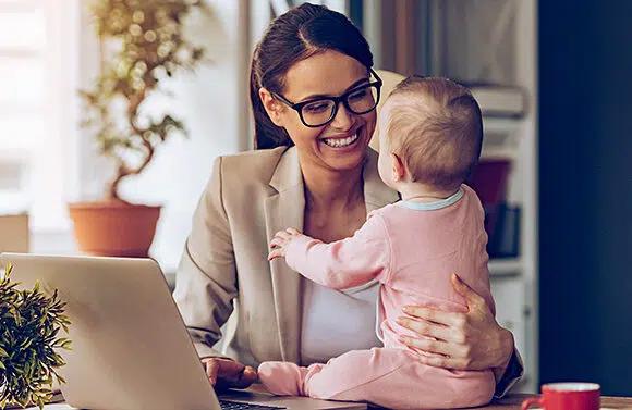 Une femme souriante tenant un bébé sur ses genoux tout en travaillant sur un ordinateur portable.