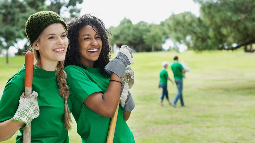 Deux jeunes femmes bénévoles souriantes, vêtues de tee-shirts verts, sapprêtent à planter dans un parc.