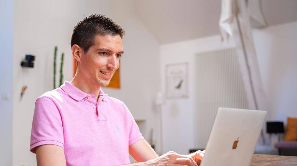 Un homme en polo rose est assis à une table et travaille sur son ordinateur portable.